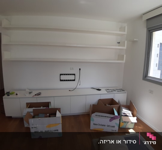 קופסאות קרטון פתוחות על רצפת עץ ליד מדפים לבנים